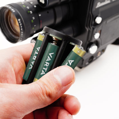 Hand hält bestückten Batterieadapter vor eine unscharfe Beaulieu Schmalfilmkamera